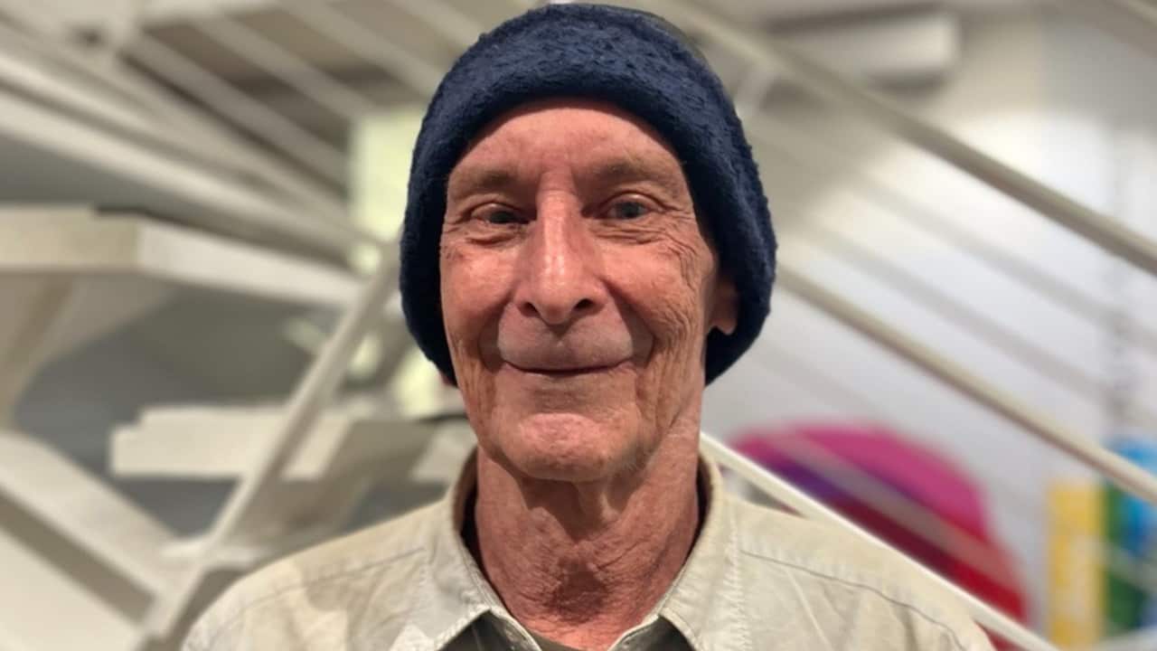 a close up of an older man wearing a blue beanie with a white office in the background