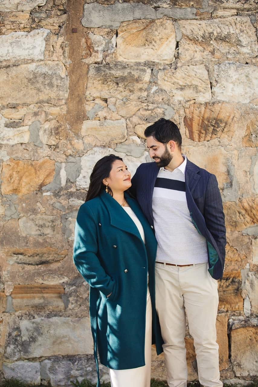 A happy couple embrace and smile at each other in front of a stone wall.