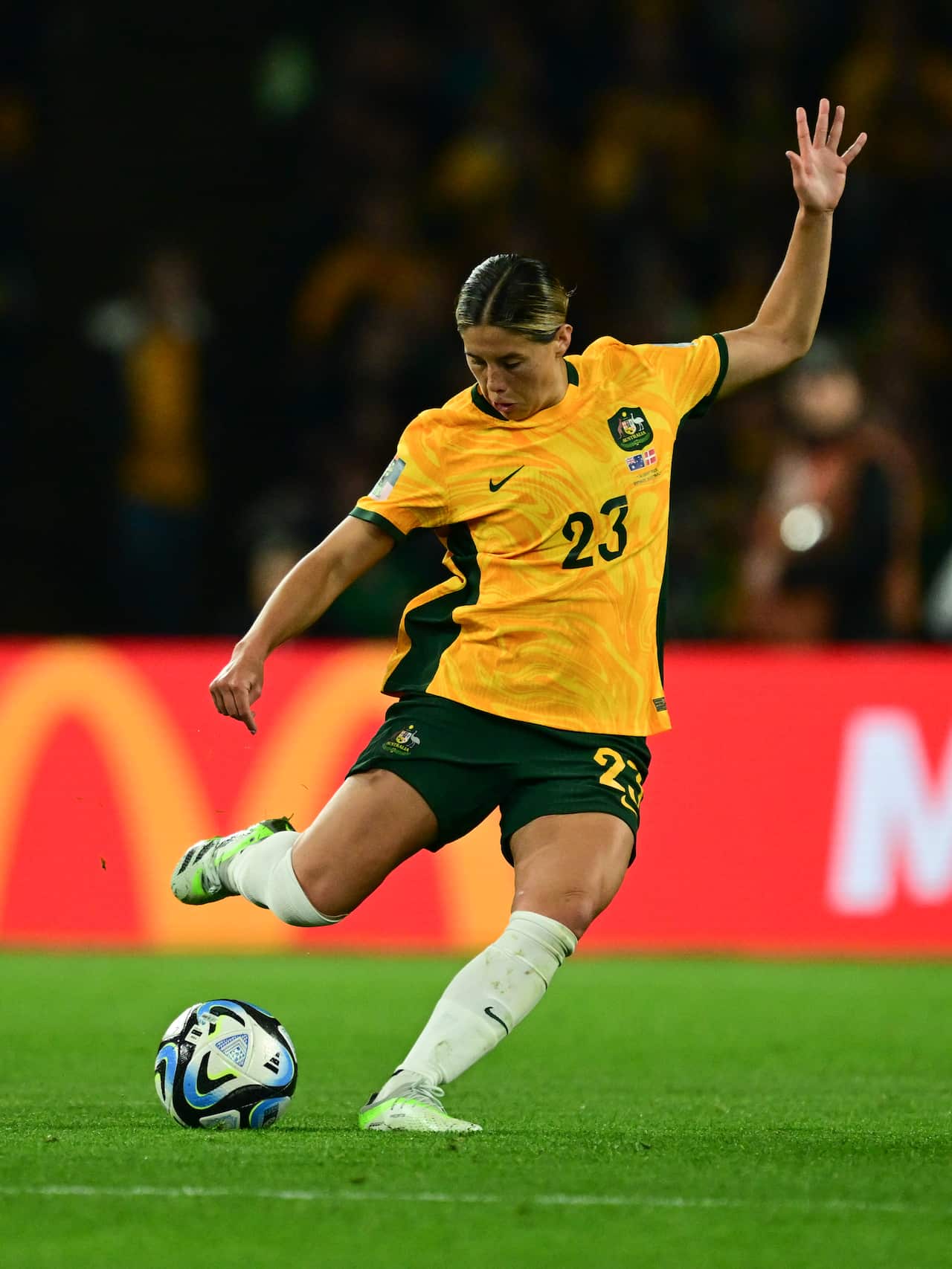A woman in a yellow football uniform kicks a ball, raising one hand above her head