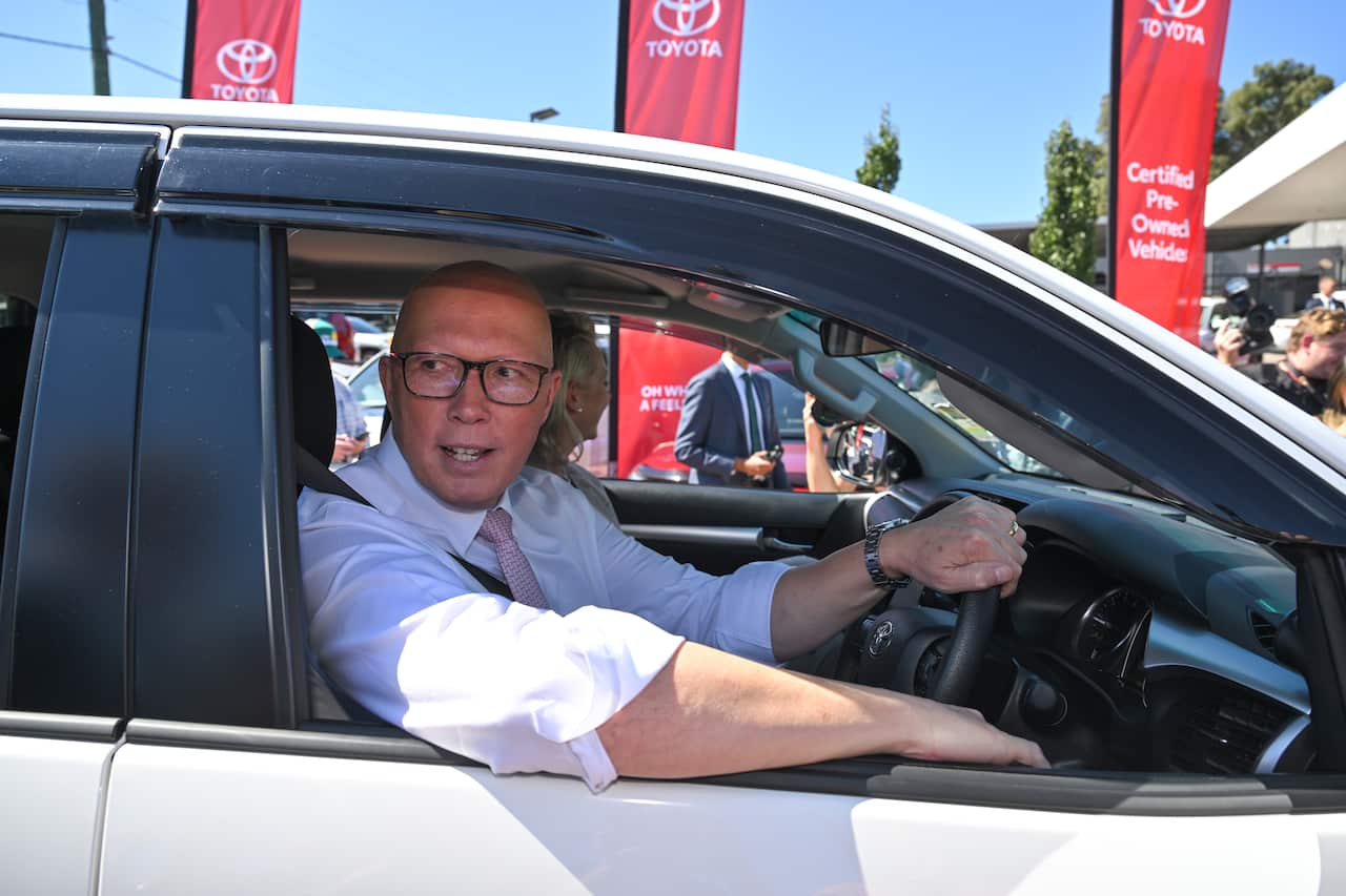 A man in a white shirt and a pink tie is sitting in the driver's seat of a vehicle with one hand on the steering wheel.