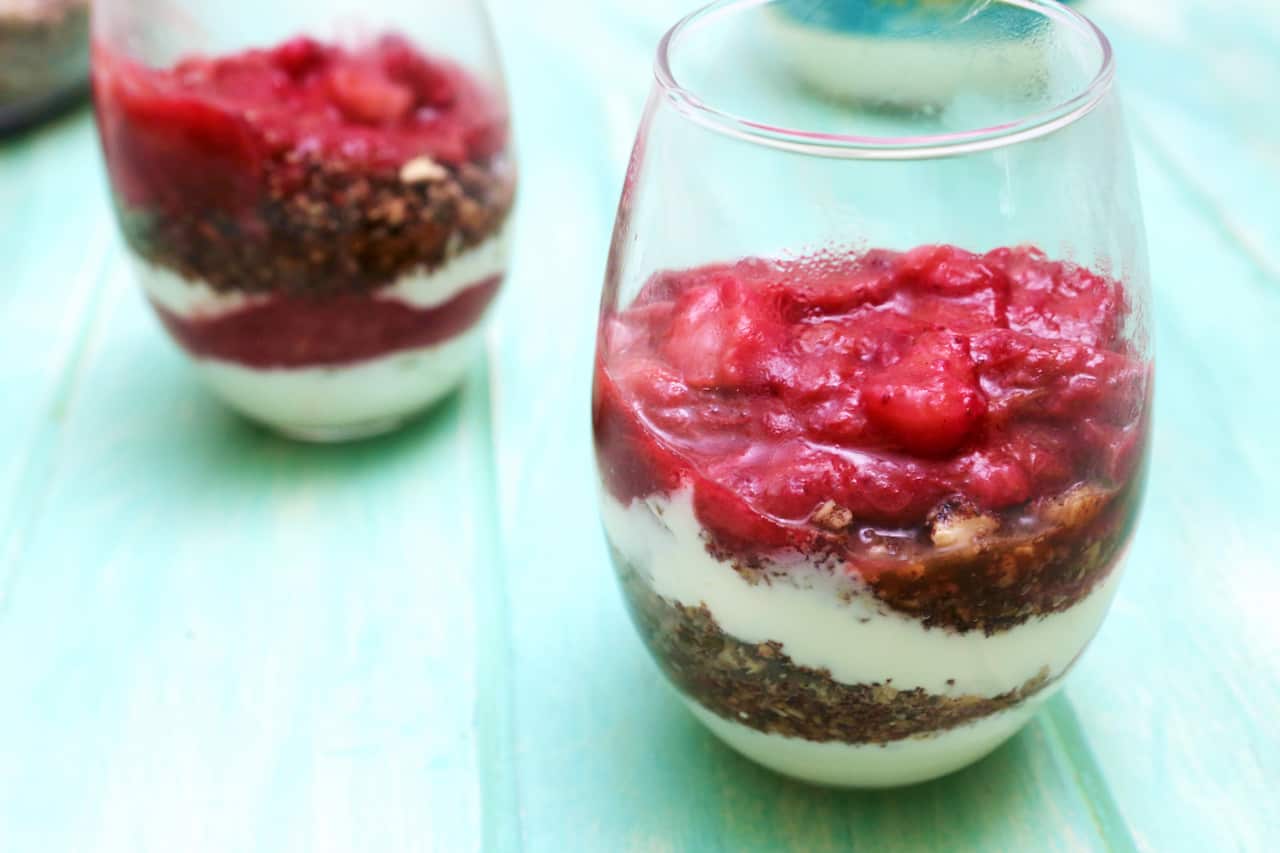 Two glasses sit on an greenish table top. Each glass holds layers of yoghurt and granola, topped with vibrant pink stewed fruit. 