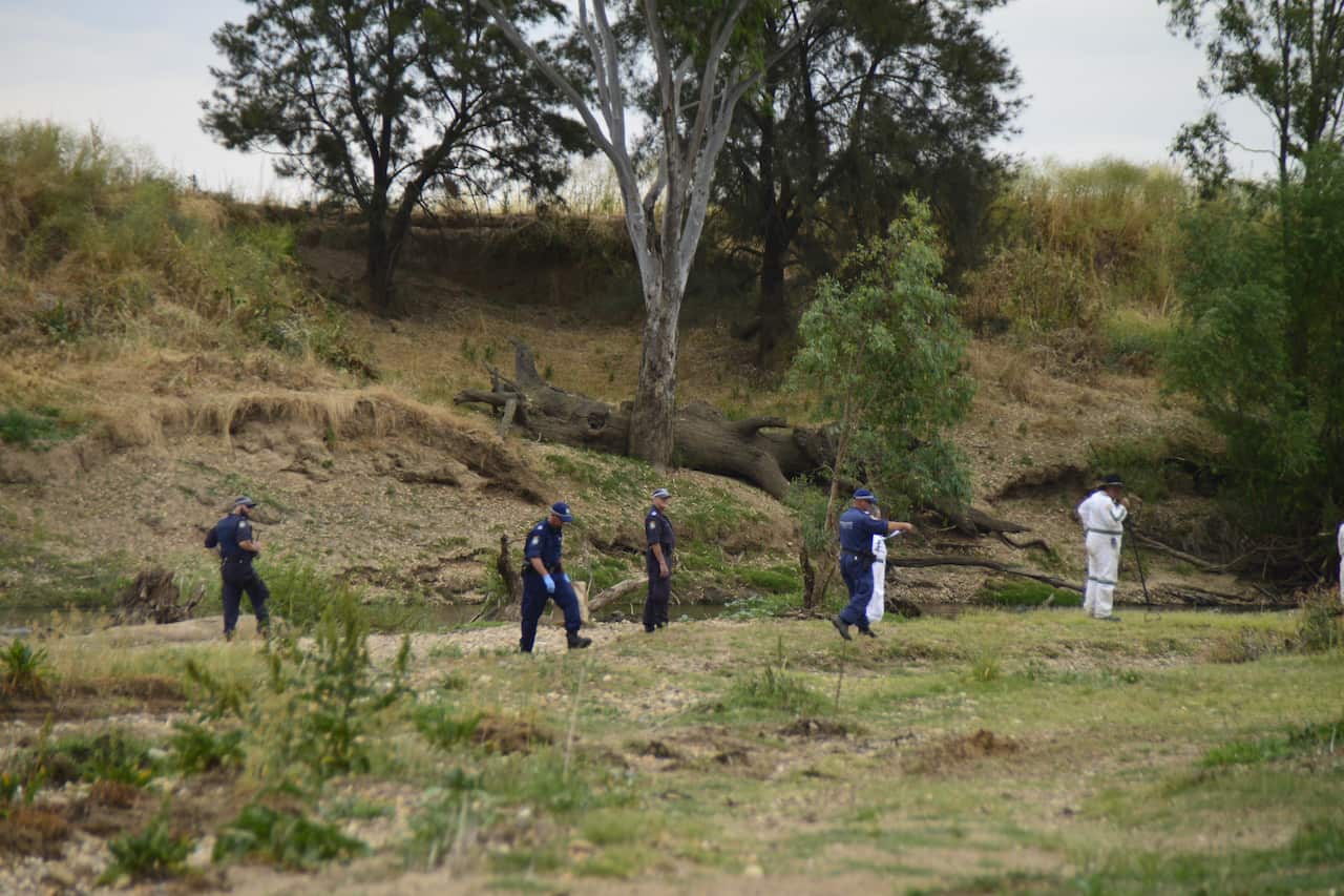 A team of five police officers are walking along a dry, grassy riverbed, with a steep, wooded bank rising behind them. The officer on the far right is wearing a white forensic suit.