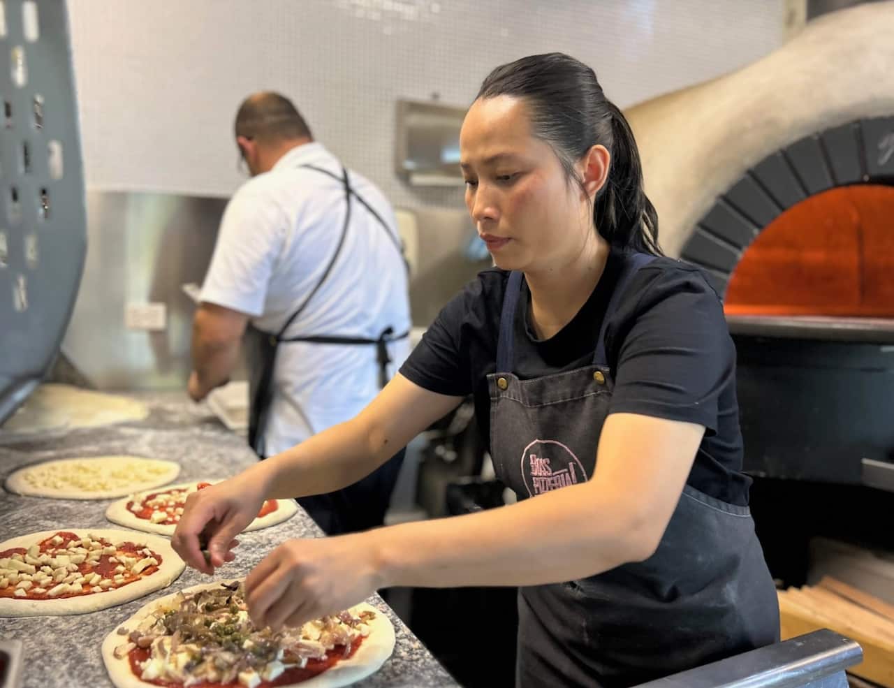 A woman in a black t-shirt stands putting topping on pizza, in front of a man in a white t-shirt with his back to camera. 