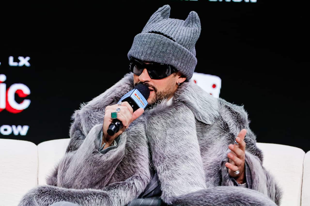 Bad Bunny holds a microphone to his mouth, sitting on a couch wearing a grey fur jacket and beanie with cat ears.