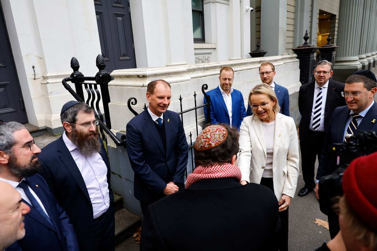 A woman in a white blazer shakes hands with a man wearing a red kippah. They are surrounded by several men.