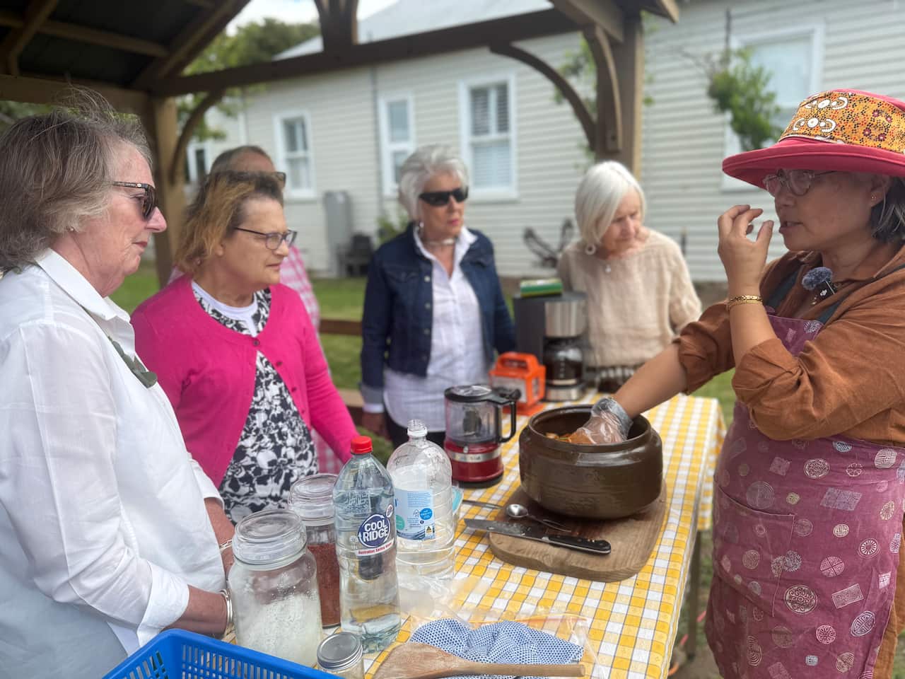 Woman explains her cooking process as five older women gather around a table watching her hands in a bowl