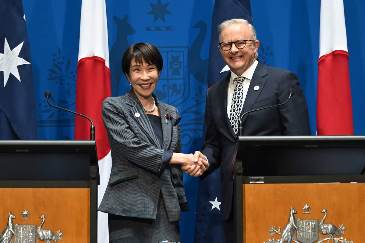 Sanae Takaichi and Anthony Albanese shaking hands. They are next to two podiums, with Australian and Japanese flags behind them.