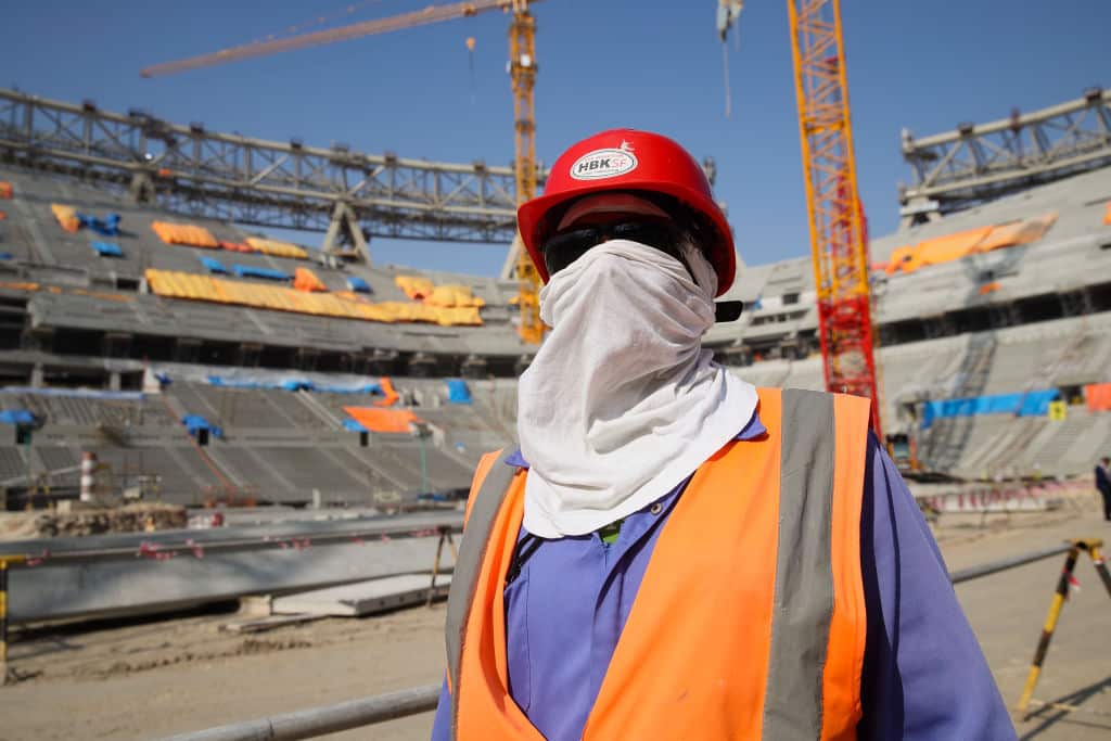 Worker standing inside stadium construction site in Qatar wearing hi-vis vest, kerchief over his face, sunglasses and hard hat