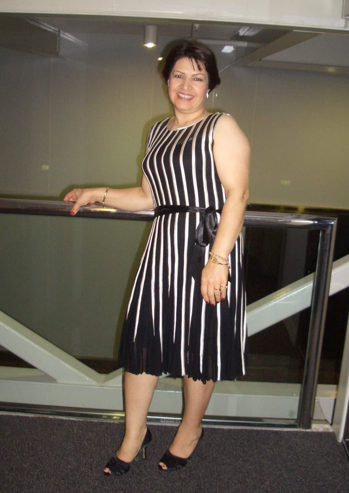 A woman in a black and white dress stands holding a railing and smiles.