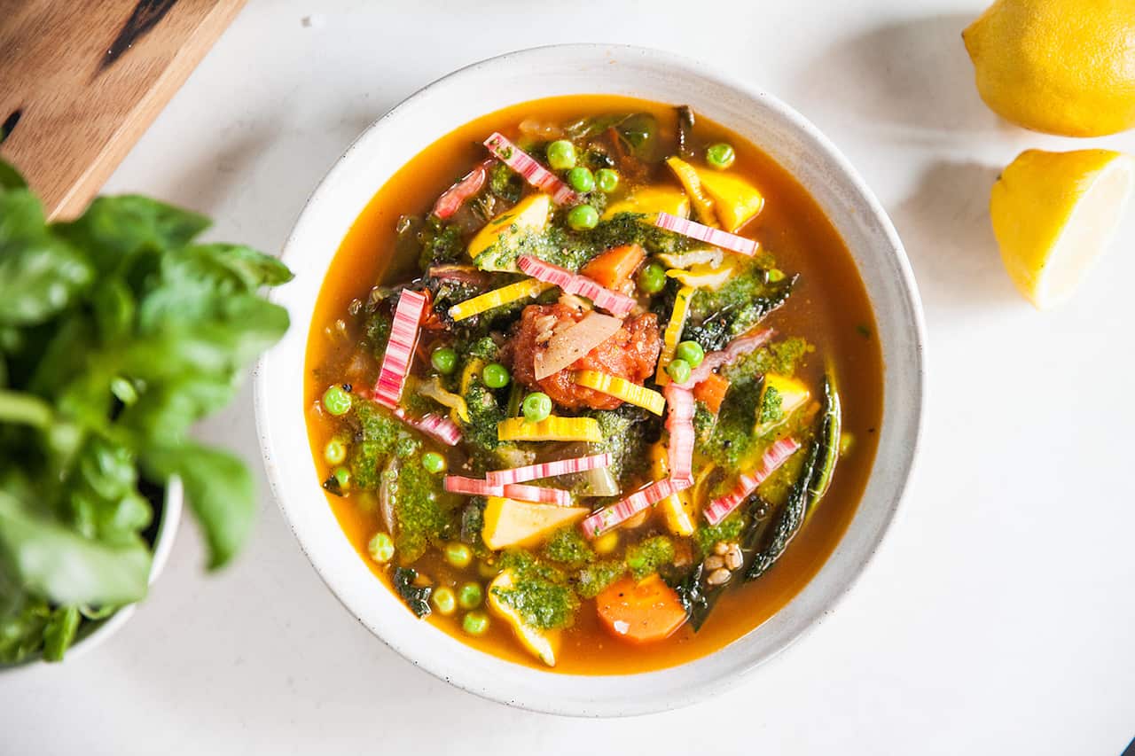 A colourful chunky vegetable soup in a white bowl, topped with a green sauce garnish, is seen from overhead. 