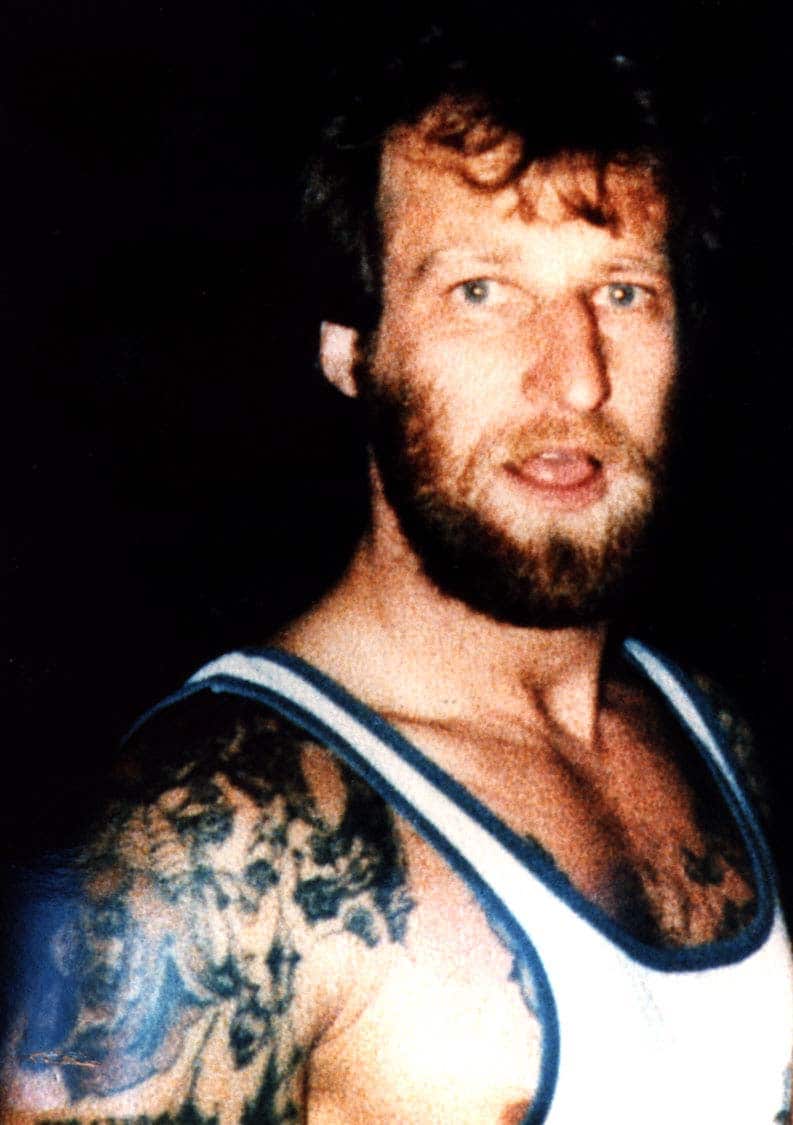 A man with a beard and tattoos on his right arm and chest wearing a white singlet with blue piping