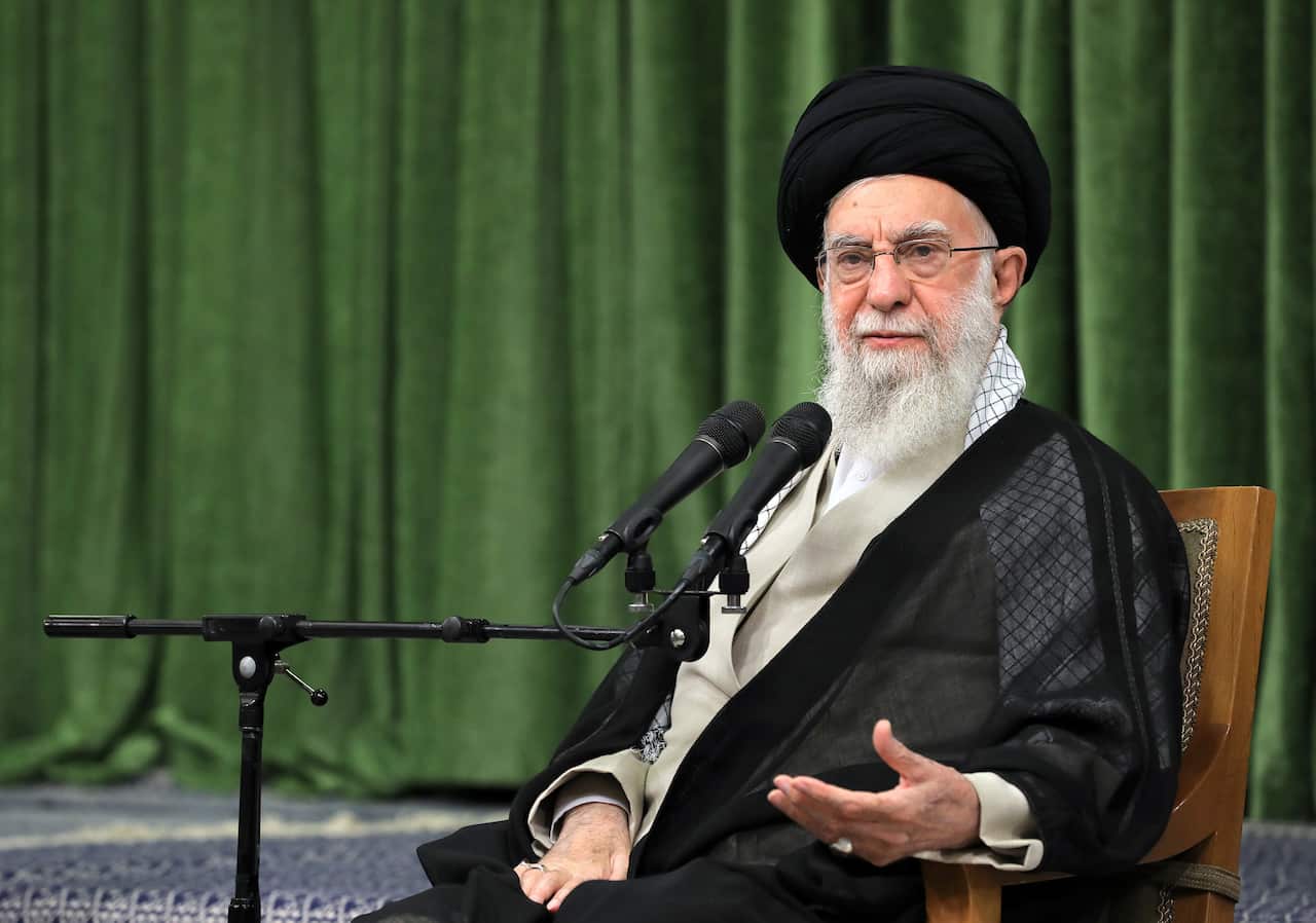 An elderly man with a long beard, wearing a black robe and turban, is sitting, speaking into a microphone, with a green curtain behind him.