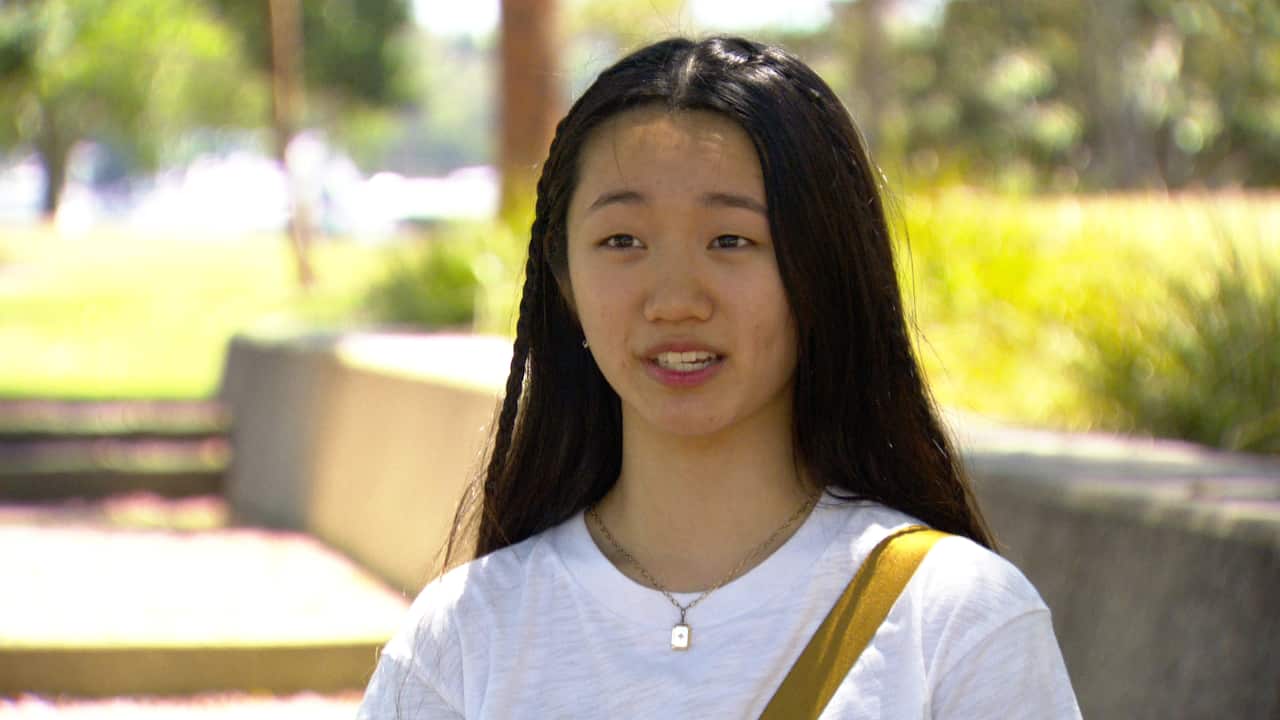 A young woman with dark hair and a white t-shirt is speaking outside