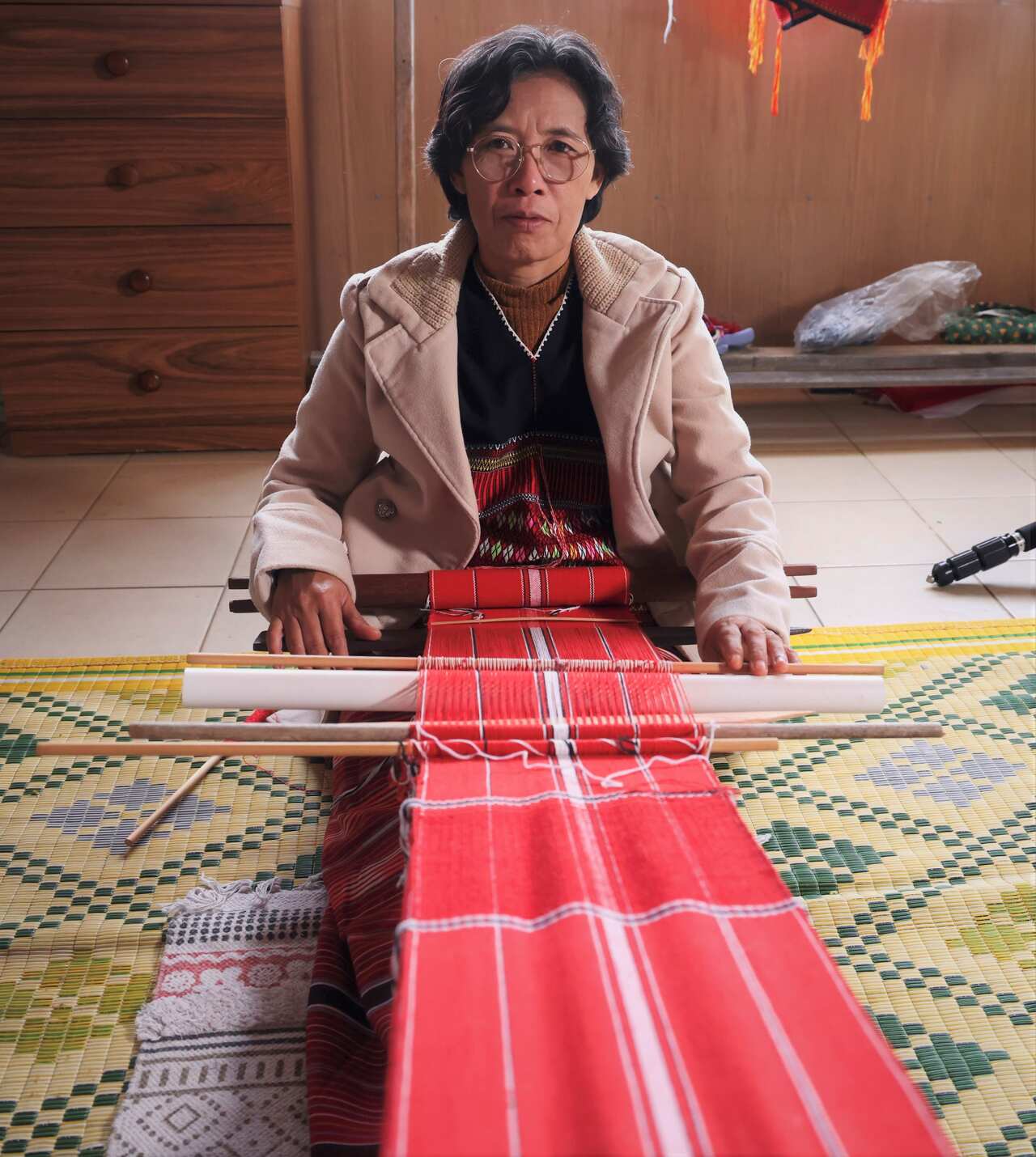 A woman in a beige jacket sits on the floor, weaving a length of red fabric on a hand-held loom. 