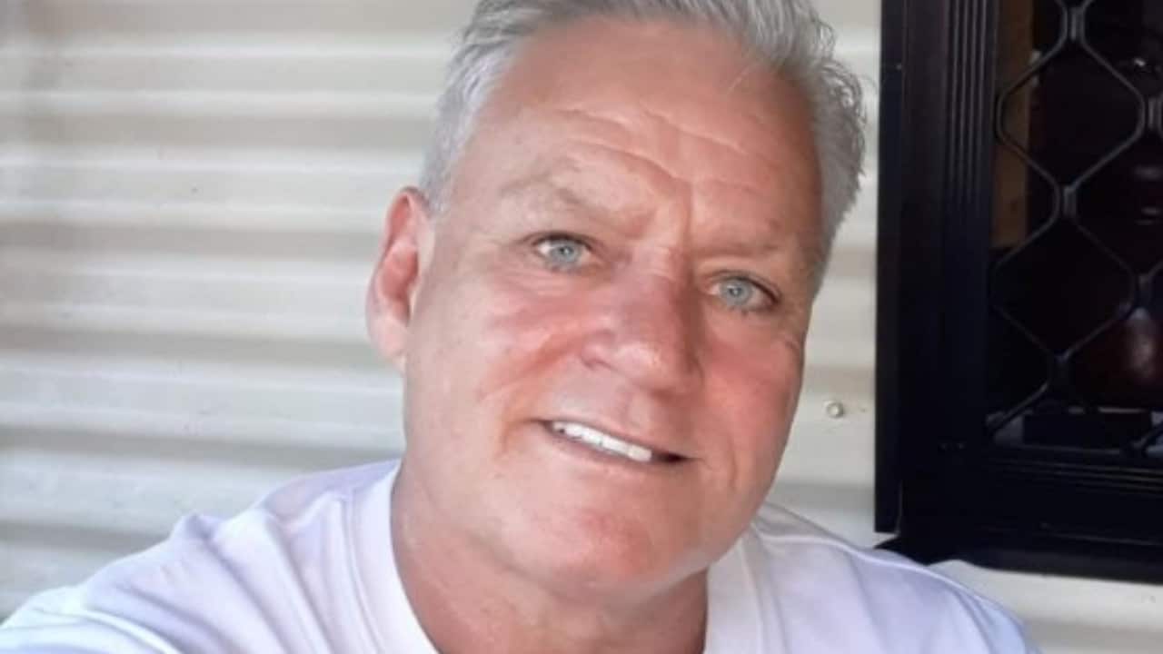 a middle aged man with tan skin and white hair smiles at the camera in front of a white background