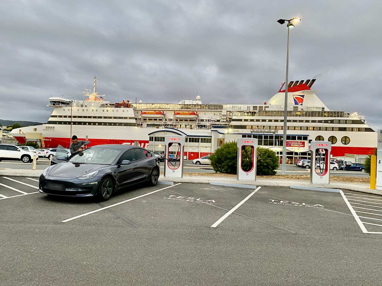 A man stands next to his car parked at a Tesla supercharger with a ferry in the background. 