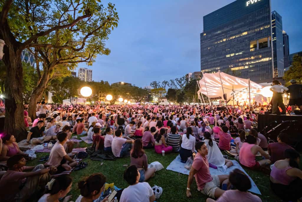 A large crowd of people mostly wearing pink sitting on grass looking at a stage