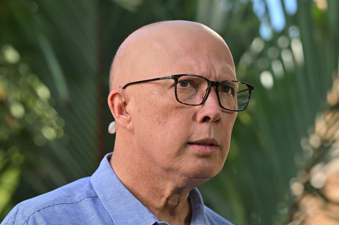 Peter Dutton wearing a blue shirt, standing in front of a green tree.