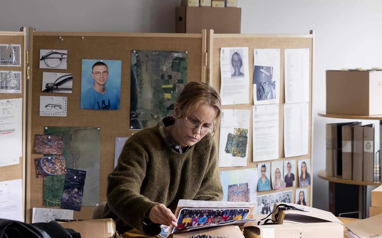 A woman with blondish, tied back hair and glasses, in a green knitted sweater, sits at a desk looking intently at a photograph. Corkboards behind her hold various pinned images and documents.