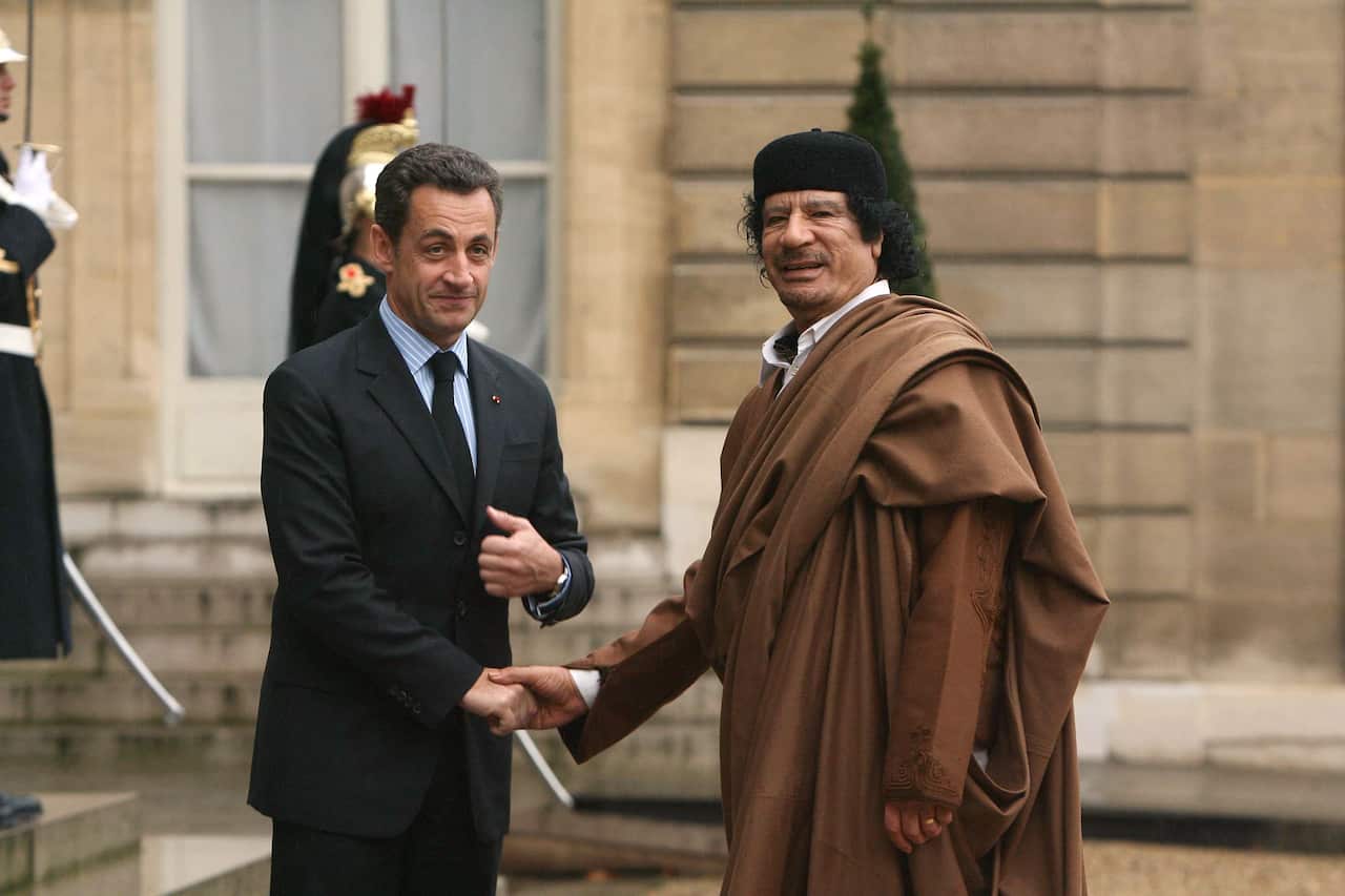 A middle-aged white man wearing a suit shakes hand with a older Arab man wearing brown robing. 