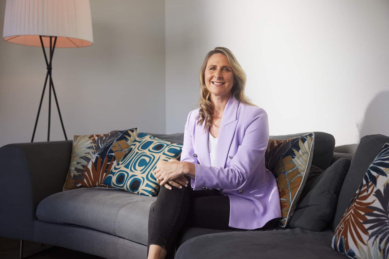 Blonde woman in a lavender blazer, smiling on a couch
