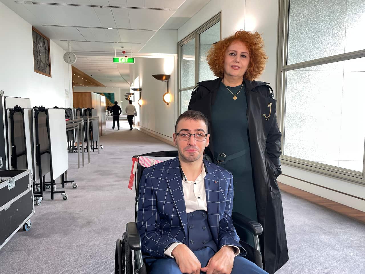 A man sitting in a wheelchair is accompanied by his mother, inside parliament house