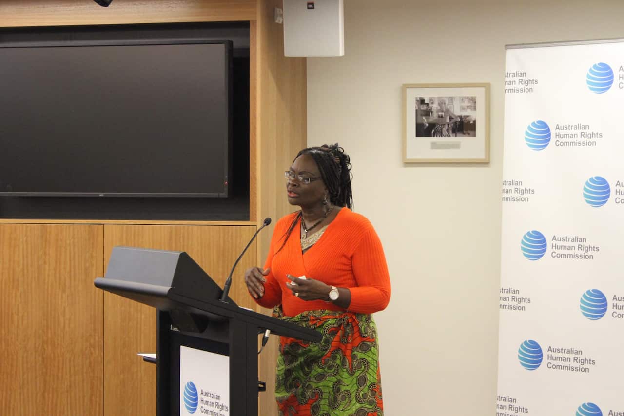 A woman wearing a bright orange top speaks at a lectern