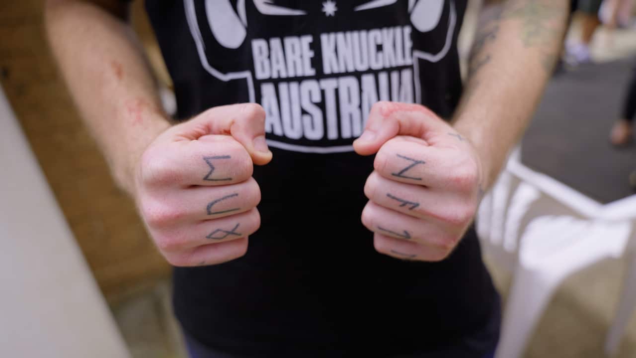 Close up of a man's tattooed fists. He is wearing a black t-shirt with white writing that reads bare knuckle Australia