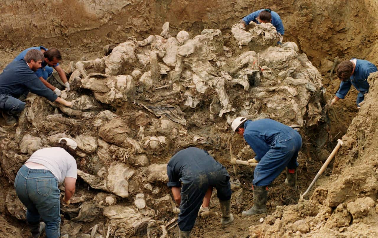 A mass grave full of dozens of decomposed bodies. Seven people wearing blue work clothes are bent over the grave site, working with tools to uncover the remains.