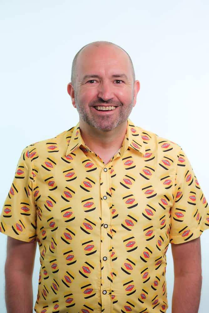 A smiling man in a collared shirt with a pattern featuring Vegemite jars.