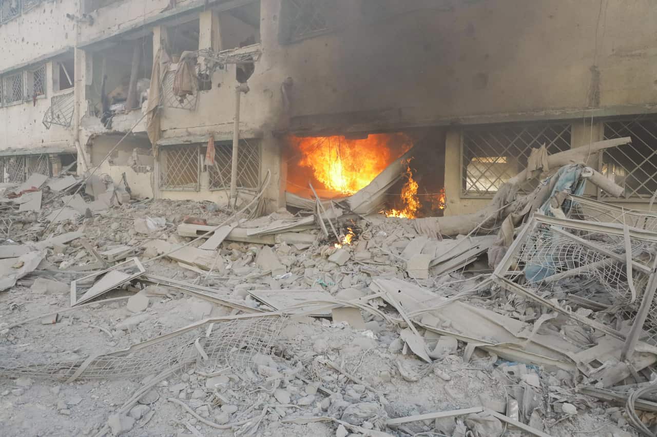A fire burns in part of a heavily damaged building covered in dust. 
