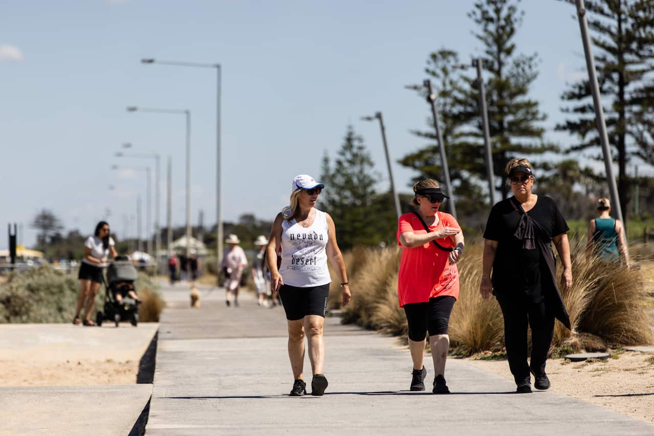 Three women in active wear and caps walk on a pavement lined by trees and sand.