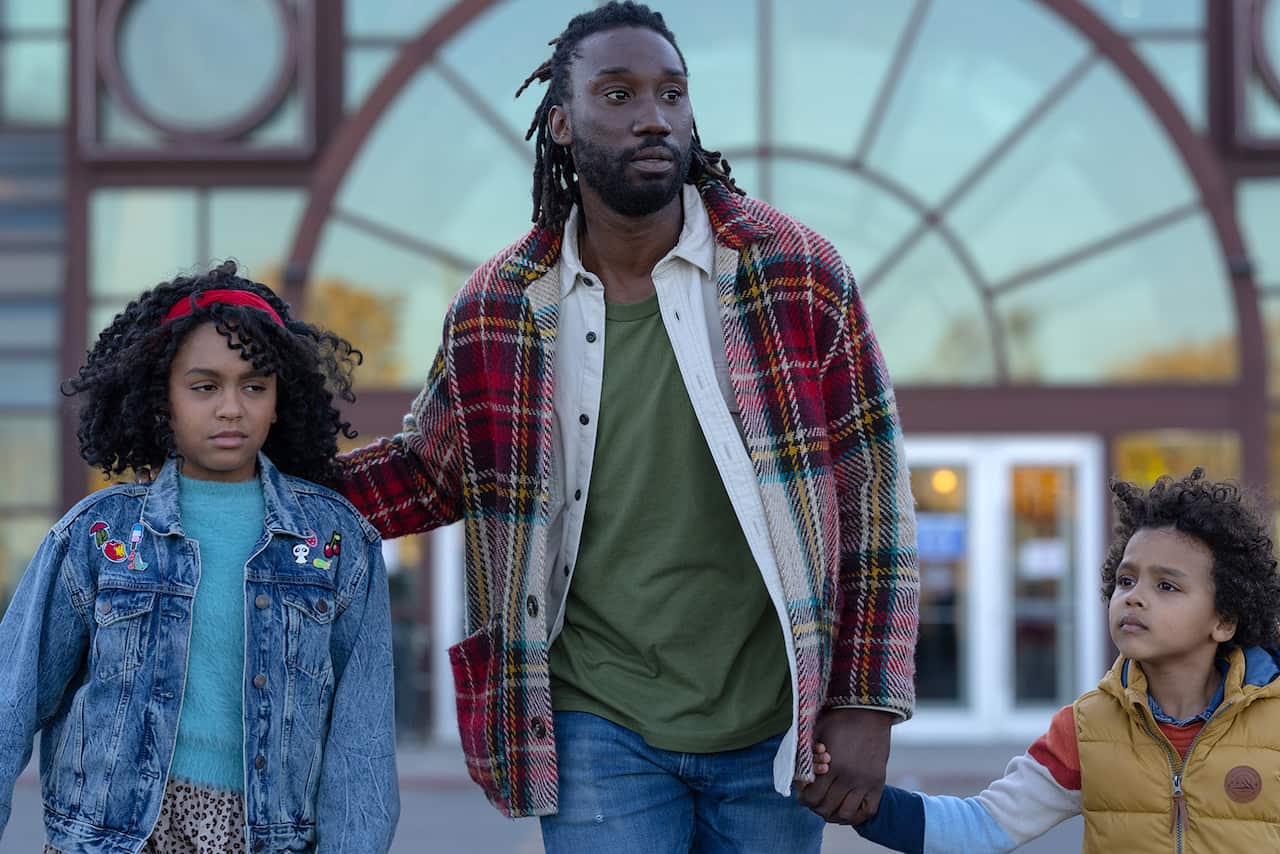 A man with dreadlocks in a green shirt and check jacket walks in front of a building, with a young girl on one side and another younger child holding his hand on the other.