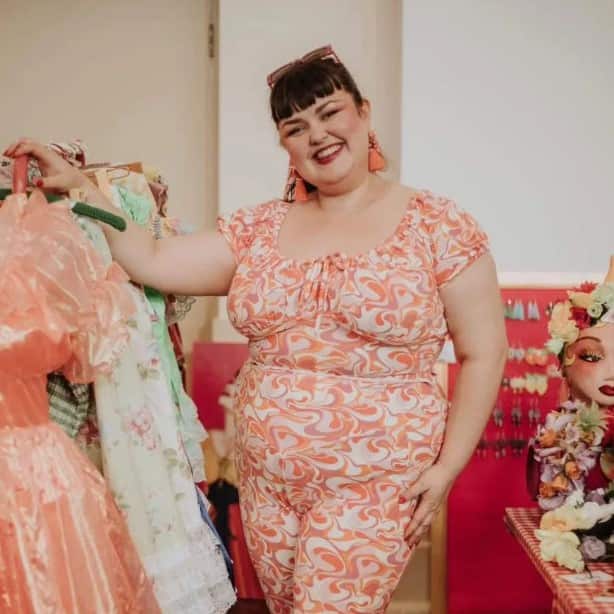 A woman with brown hair and white skin stands next to a clothing rack. She's wearing clothes with a pink, white and orange swirl pattern.