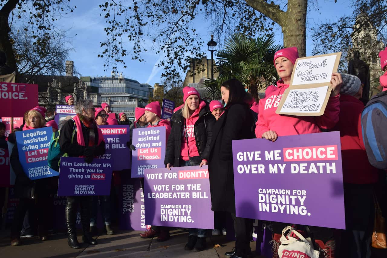 A group of people wearing pink and holding purple signs in support of assisted dying.