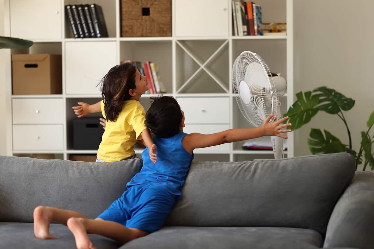 Children are spreading their arms in front of a standing fan inside a house.