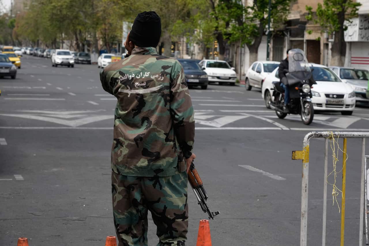 An armed individual in camouflage uniform and a black beanie stands at a street checkpoint, seen from behind with Persian text and a sword emblem on their jacket.