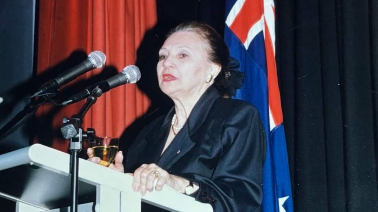 an older woman in a black silk suit speaking at a lectern with an Australian flag behind her