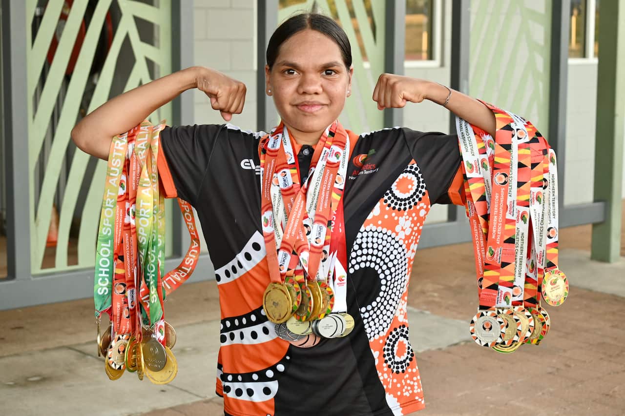 A young woman flexes her arms. She has many medals hung on each arm and around her neck