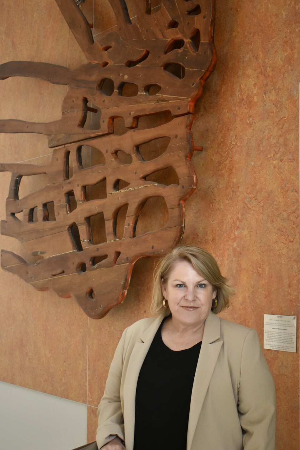 A woman in a suit stands against a brown background with a neutral expression on her face.