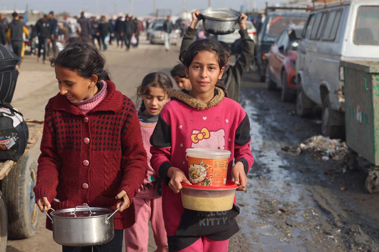 Three young girls dressed in pink and red carry food rations along a busy road.