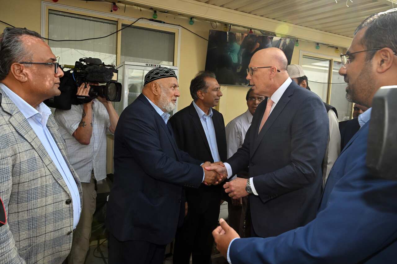 A man in a dark suit shakes hands with another person, also in a dark suit and wearing a skull cap, while men and camerapersons stand around them, observing the exchange.