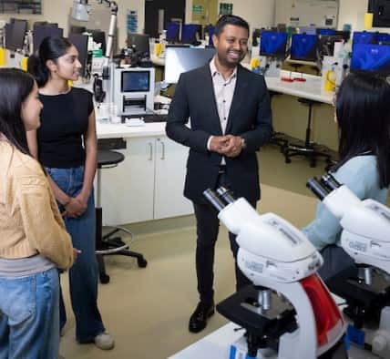 A man in a black suit stands in a science lab, talking to three students. 
