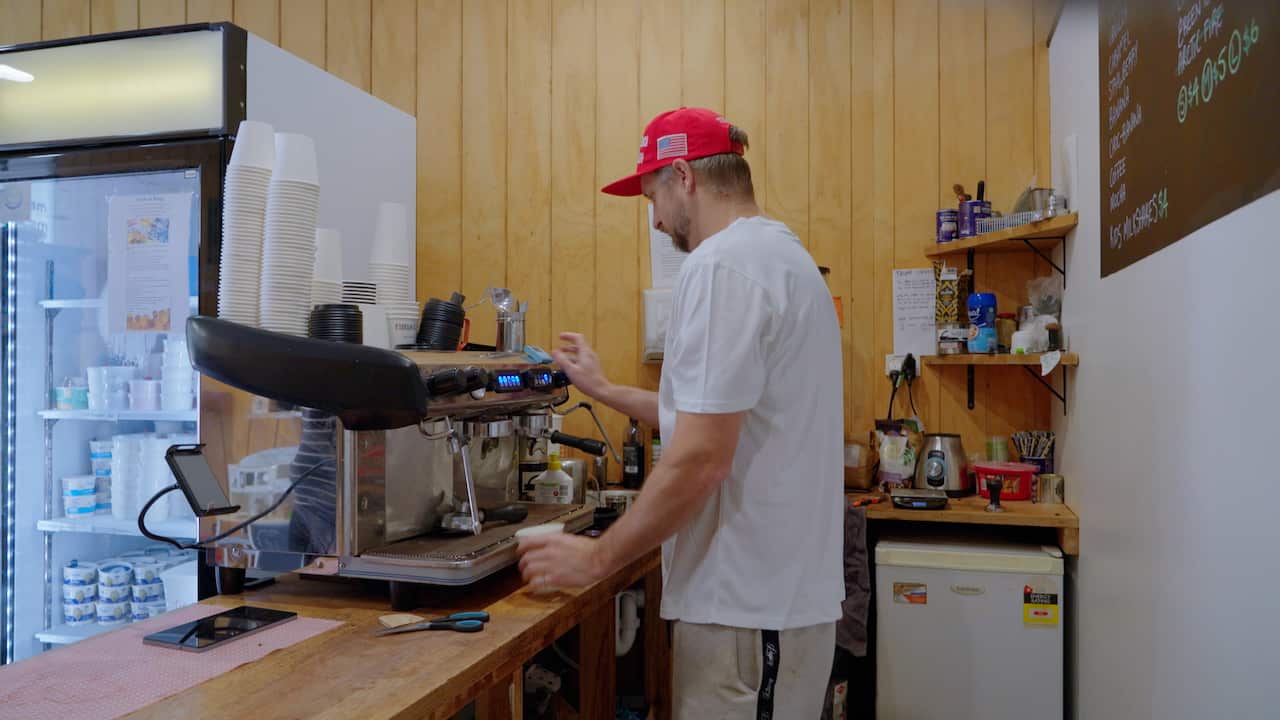 A man wearing a white t shirt is making a coffee while wearing a Make America Great Again hat