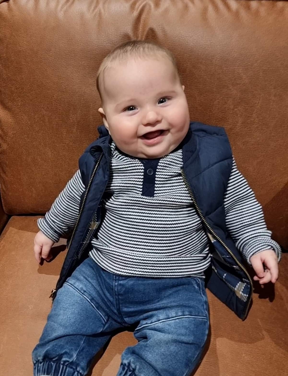 A smiling baby wearing blue jeans, a blue striped shirt and a blue sleeveless puffer jacket sitting on a brown couch.