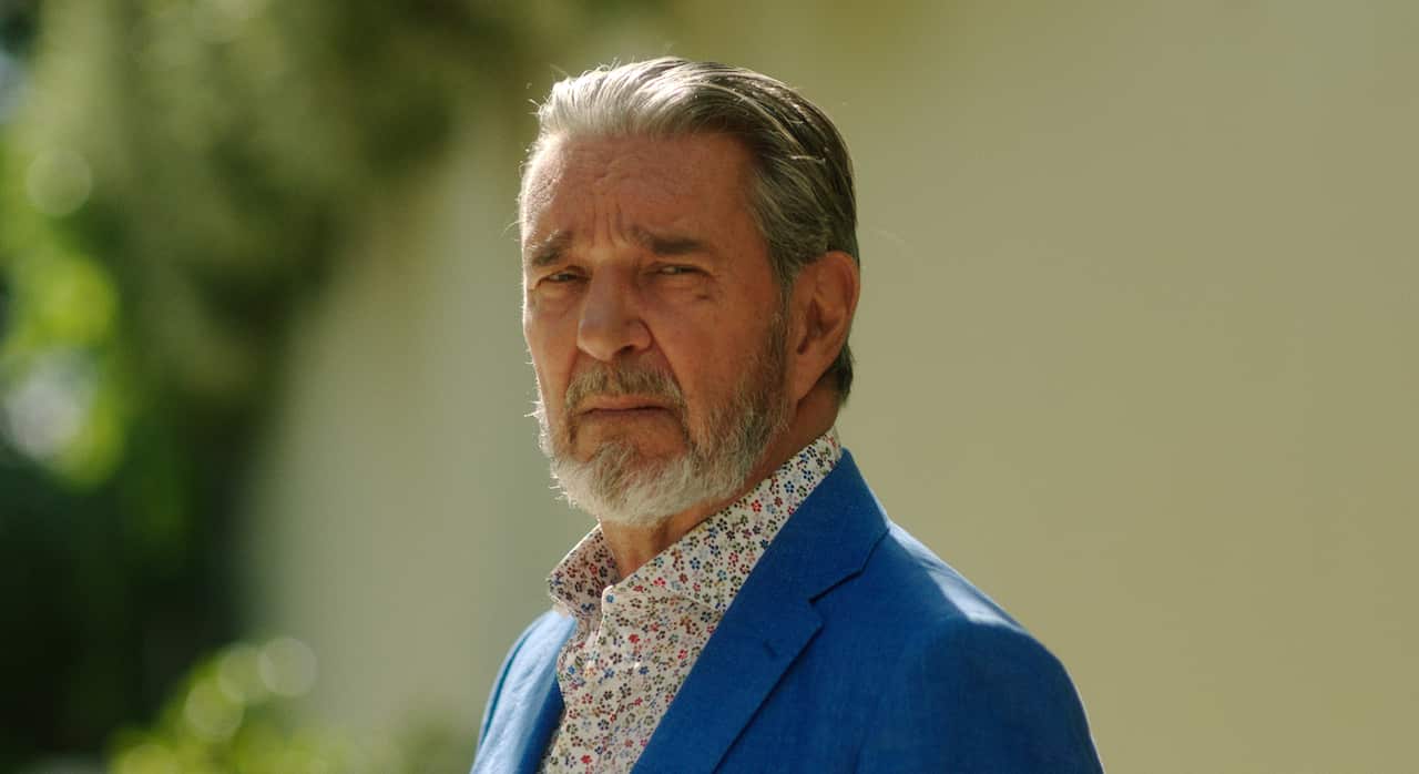 A well-dressed grey-haired man in a blue jacket looks sideways.