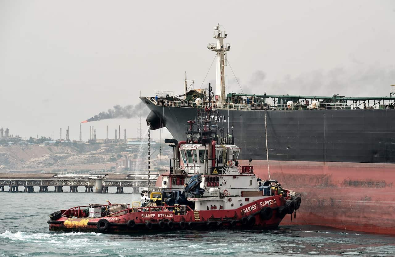 A smaller boat alongside an oil tanker at a sea port with an oil refinery in the background