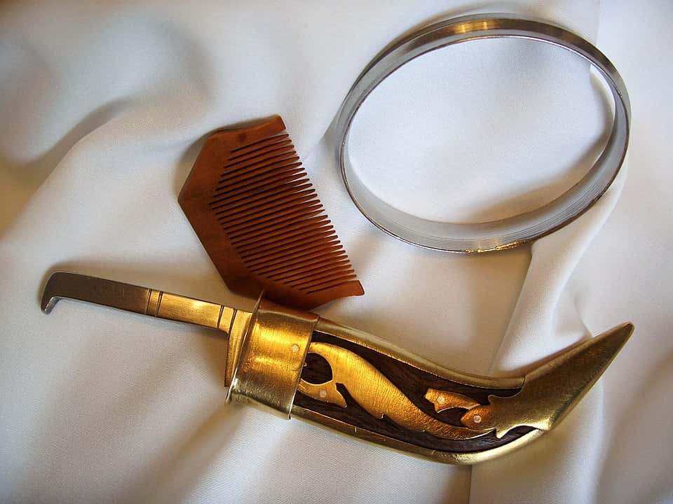 A close-up photograph of three of the five Sikh articles of faith resting on a white cloth: a wooden comb (Kanga), a curved, brass-hilted miniature sword (Kirpan), and a silver metal bracelet (Kara).