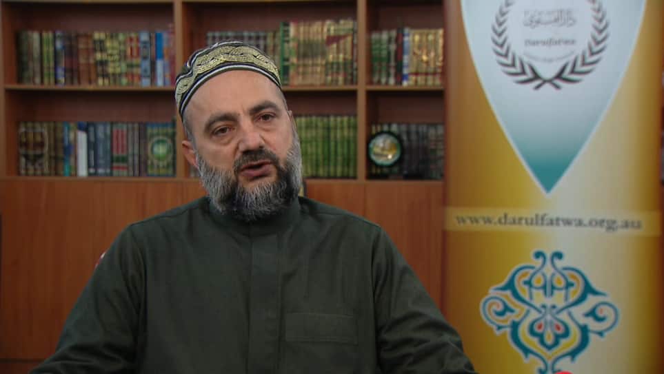 A man wearing an embroidered skull cap and a grey Arabic robe sits in an office, with a Darulfatwa banner and a bookshelf in the background.