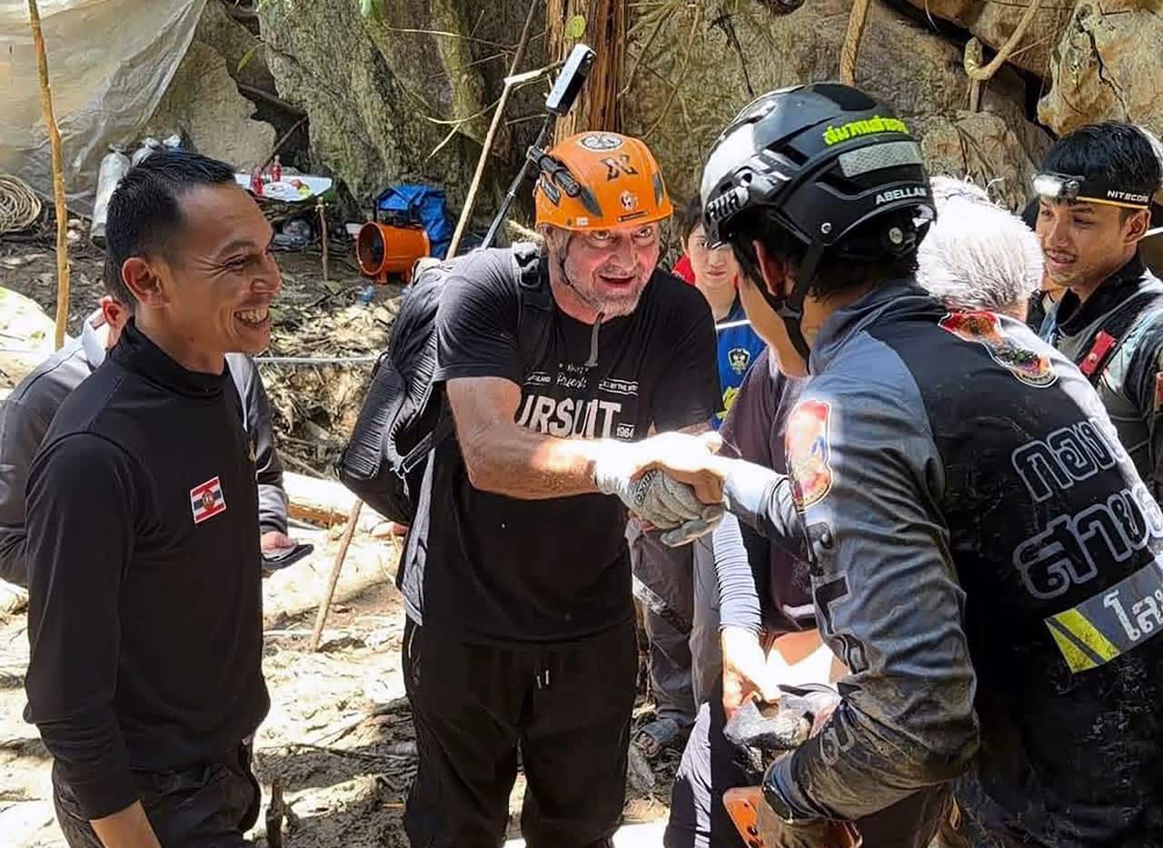 A middle-aged white man in rescue gear shakes the hand of a Thai rescuer in front of muddy boulders.